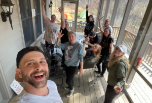 Eight smiling people raise their arms in a celebratory fashion standing on a screen-enclosed sun porch. 