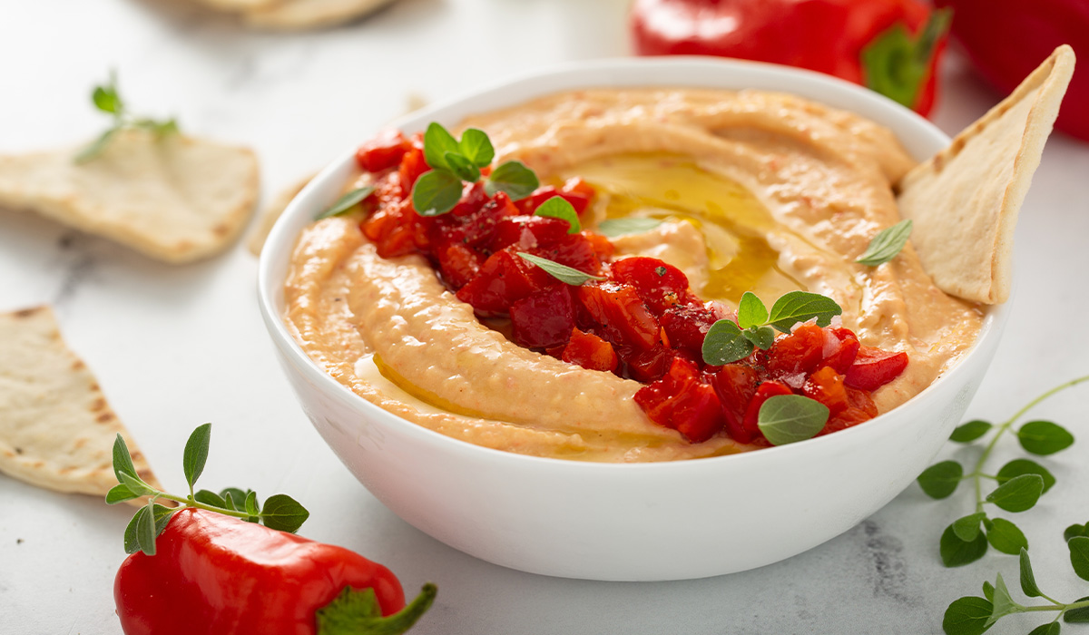 Roasted hummus with red pepper garnish in a white bowl, surrounded by whole ingredients. 