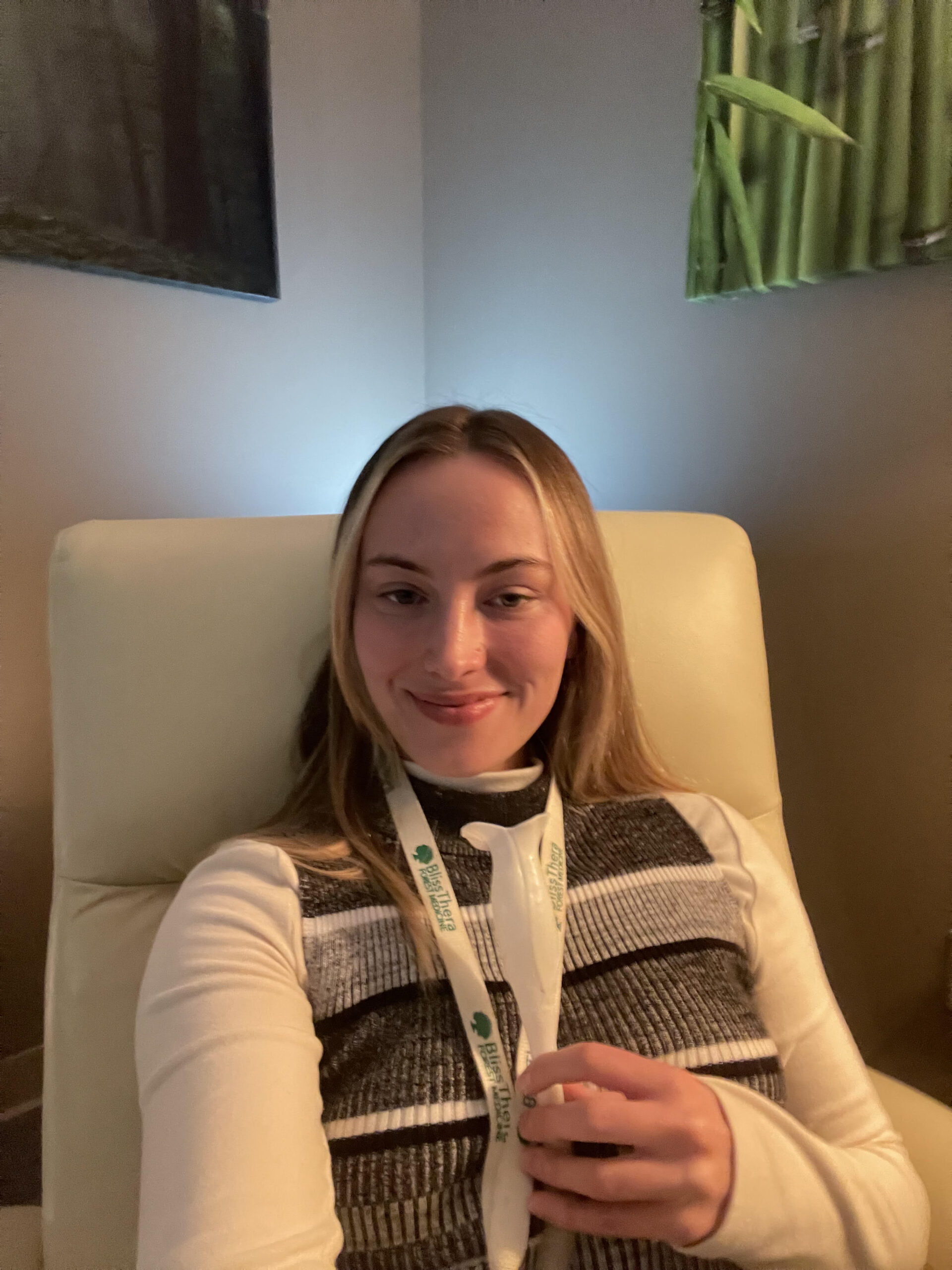 A woman in a striped shirt sits in a white armchair and holds the breathing tube attached to the BlissThera device.